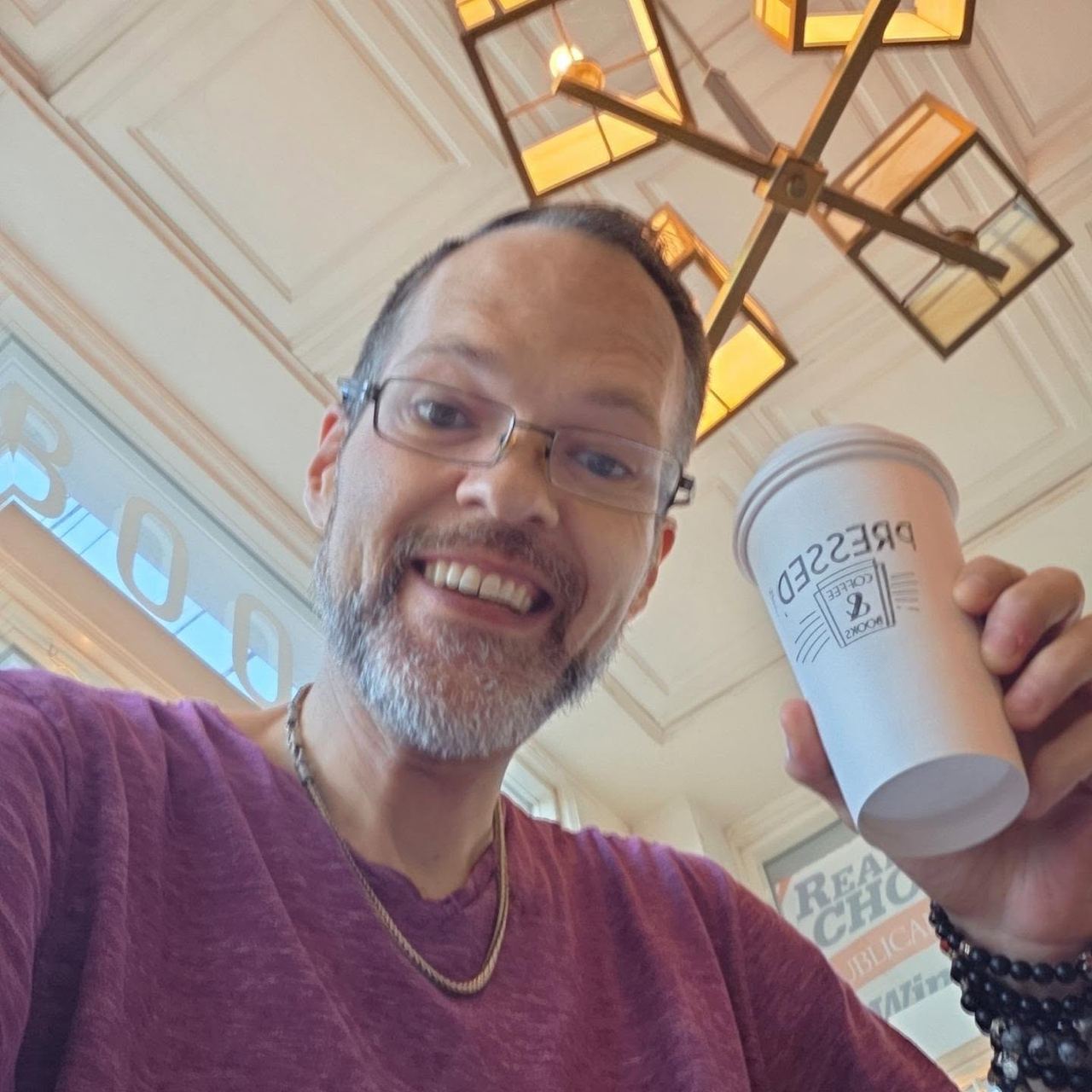 David Strausser grinning in a selfie, holding a Pressed Coffee and Books to-go cup under a geometric pendant light