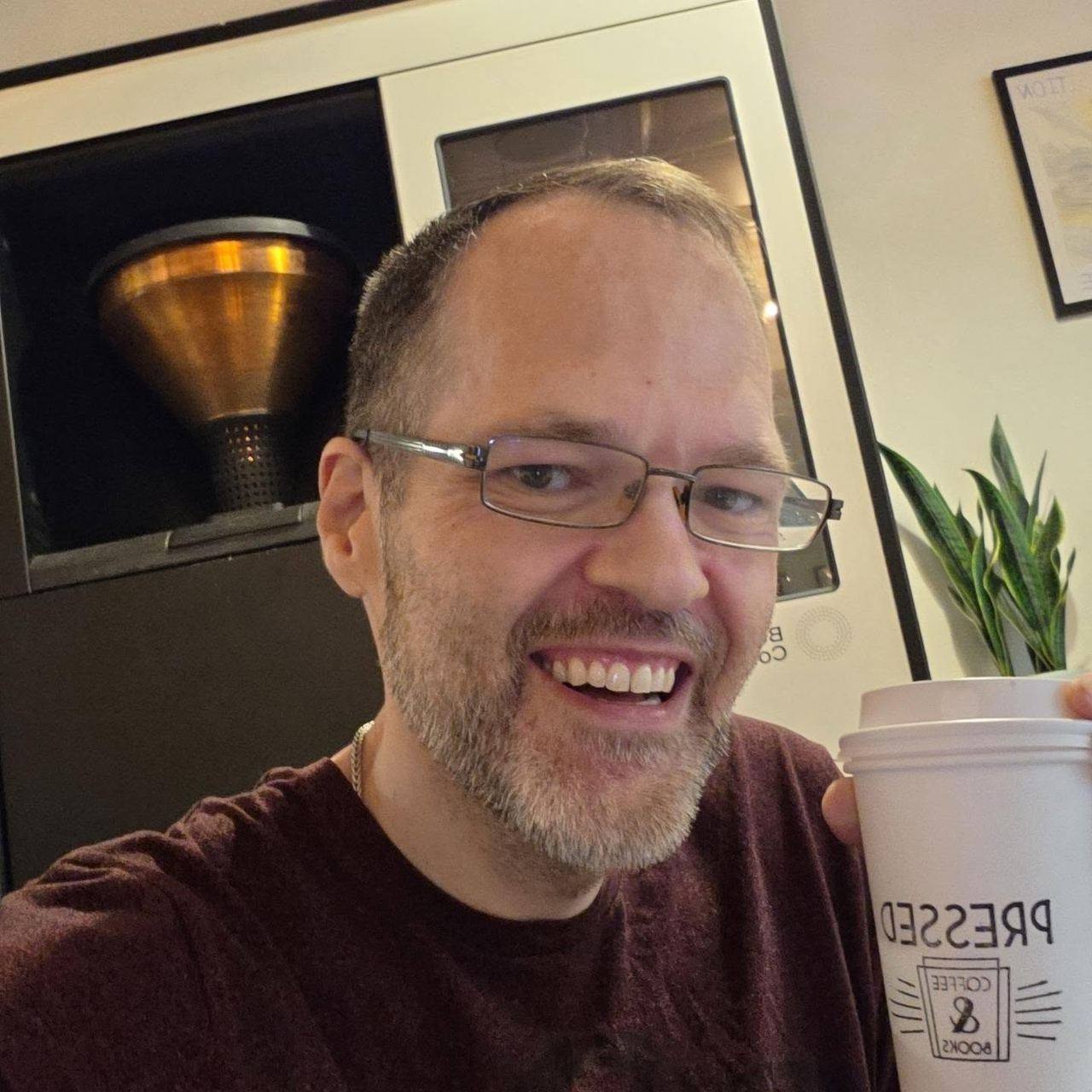David Strausser smiling with a Pressed Coffee and Books cup, a copper coffee machine behind him